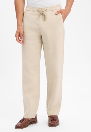 Mann trägt beige Leinenshosen mit Kordelzug und Seitentaschen sowie braune Lederloafers mit Quasten, steht vor einem weißen Hintergrund.