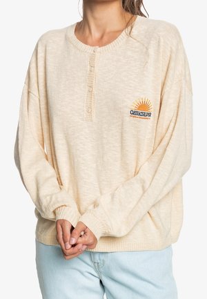 Mujer con un jersey de punto beige Quiksilver con cuello abotonado y vaqueros azul claro, con las manos entrelazadas al frente.