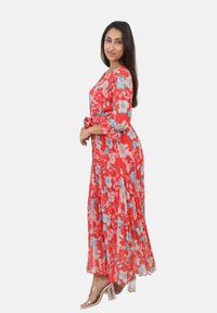 Robe maxi florale rouge avec des accents bleus, dotée d'un décolleté en V, de manches ¾ et d'une taille à nouer. Un tissu transparent ajoute de la texture et du mouvement. Sandales à talons transparents.