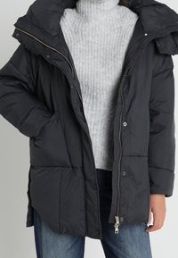 Černá vatovaná bunda s předním zapínáním na zip a druky, nadměrnou kapucí a prošívaným vzorem, nošená přes světle šedý žebrovaný svetr.