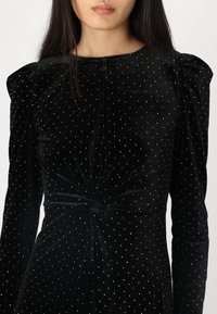 Sorte fløyelskjole med vridd front, lange puffermer og små sølvprikker over hele stoffet.