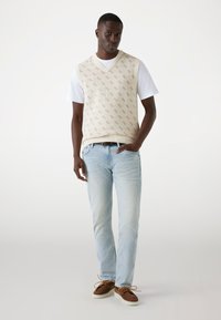 Beige gestrickte Weste mit Rautenmuster über einem weißen T-Shirt, kombiniert mit hellblauen Jeans und braunen Schnürschuhen.
