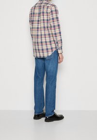 Man die blauwe, recht gesneden spijkerbroek, zwarte leren schoenen en een geruite blouse met lange mouwen draagt, staand tegen een effen witte achtergrond.