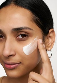 Une crème blanche est appliquée sur la joue par une main. La peau est lisse avec une finition naturelle. Les cheveux sont noirs et tirés en arrière.