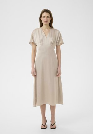 Kobieta stojąca w beżowej sukience midi z krótkimi koronkowymi rękawami i dekoltem w serek, w połączeniu z czarnymi sandałami na ramiączkach, na jednolitym jasnym tle.