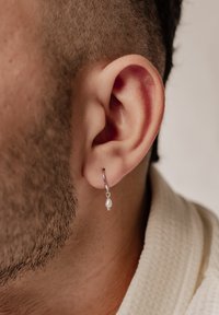 Orecchino ad anello in argento con un piccolo pendente di perla, indossato all'orecchio. Il design include una chiusura semplice e una finitura metallica liscia.
