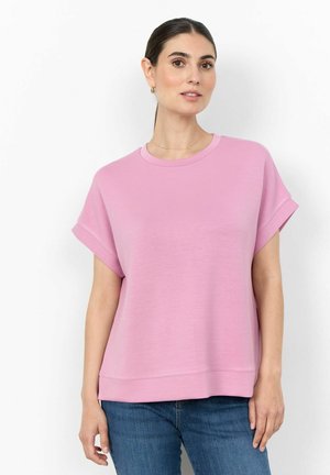 Kvinde med mørkt hår bundet tilbage iført en løs, kortærmet lyserød top og blå jeans, stående mod en hvid baggrund.