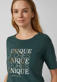 Donkergroene katoenen t-shirt met korte mouwen, voorzien van een voorprint van de woorden "UNIEKE VERHALEN" in verschillende lettergroottes en kleuren.