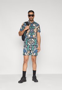 Πουκάμισο με κοντά μανίκια και μοτίβα, καθώς και σορτς που ταιριάζουν σε σκούρο ύφασμα με πολύχρωμα αφηρημένα σχέδια, συνδυασμένα με μαύρο σακίδιο και γυαλιά ηλίου.