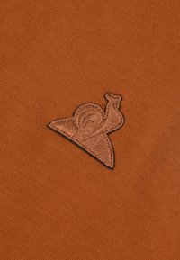 Sfondo in tessuto marrone con un logo a forma di lumaca cucito in nero, caratterizzato da un design a spirale del guscio, posizionato nell'angolo inferiore.