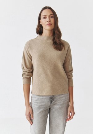 Pull beige à manches longues avec un col haut, texture douce et coupe décontractée. Assorti à un jean gris clair, présentant des éléments de design simples.