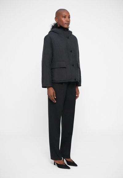 Giubbotto imbottito nero con cappuccio foderato di pelliccia, bottoni frontali e due grandi tasche applicate, indossato sopra pantaloni neri sartoriali e tacchi.