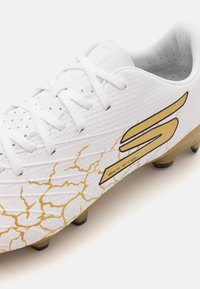 Bottes de football blanches avec une surface texturée, présentant des détails en or craquelé et un logo bien visible. Conception légère avec des crampons noirs.