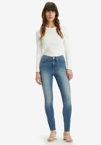 Jeans denim biru ramping dengan sentuhan pudar, lima saku, dan pinggang tinggi, dipadukan dengan atasan putih lengan panjang dan sepatu hak tinggi bersandel.