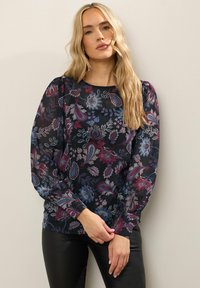 Blusa floreale multicolore con maniche lunghe, scollatura a barca e sfondo scuro; presenta motivi blu, rossi e bianchi su un tessuto trasparente.