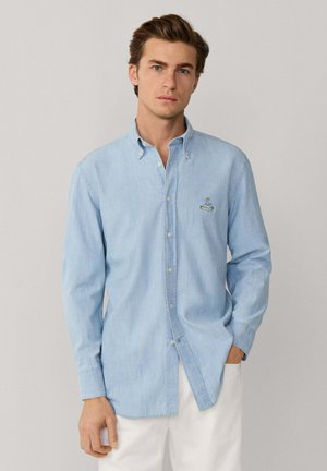 Jeune homme portant une chemise bleue clair à boutons avec manches longues et un pantalon blanc, debout devant un fond clair uni.