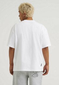 Biała bawełniana koszulka z krótkim rękawem, luźny krój i mały haftowany detal na dolnym brzegu. Gładki tylny widok.