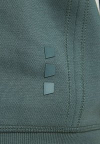Tessuto verde scuro con una trama strutturata, caratterizzato da tre piccoli quadrati in una tonalità abbinata, posizionati verticalmente sulla cucitura laterale.