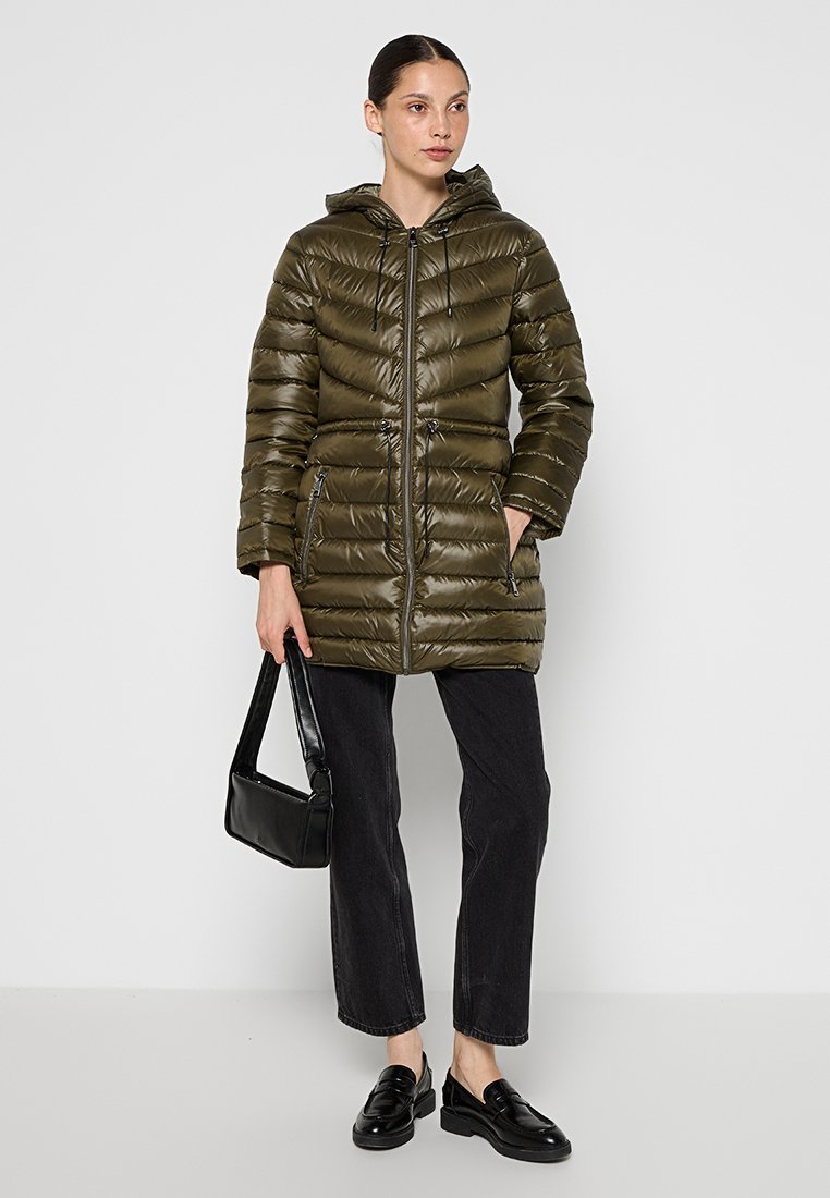 Abrigo de plumas verde oliva con capucha, que cuenta con un cierre frontal, costuras horizontales y dos bolsillos laterales. Combinado con pantalones negros y mocasines.