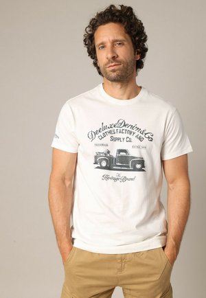 Homme aux cheveux bouclés portant un t-shirt graphique blanc avec un camion vintage et un pantalon khaki, debout les mains dans les poches.