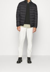 Zwarte pufferjas met horizontale stiksels, gedragen over een donkergroene trui, licht overhemd met kraag, witte broek en zwarte enkellaarzen.