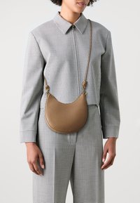 Borsa a tracolla in pelle marrone con forma a mezzaluna, texture a granuli e tracolla regolabile, indossata su un outfit a fantasia check.