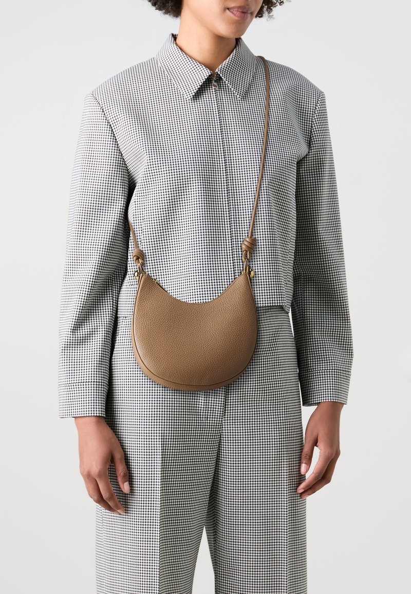 Borsa a tracolla in pelle marrone con forma a mezzaluna, texture a granuli e tracolla regolabile, indossata su un outfit a fantasia check.
