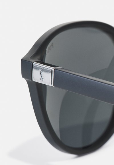 Melna saulesbrilles ar noapaļotu dizainu, pelēkajiem tonētajiem stikliem un sudraba logotipa detaļu uz kājiņas. Gluda, matēta tekstūra.