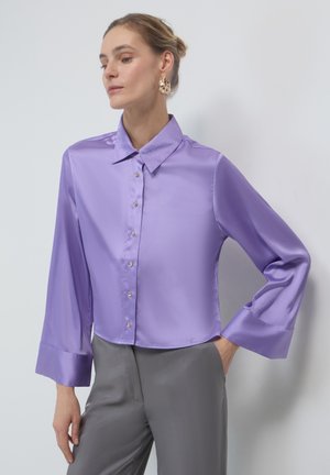 Lavendelfarbene Satinbluse mit klassischem Kragen, langen Glockenärmeln und vorderem Knopfverschluss. Kombiniert mit grauen Satin-Hosen.