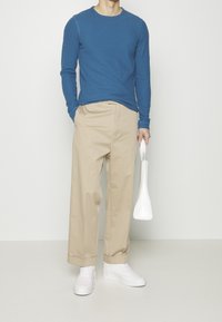 Blå långärmad skjorta, beige vida byxor, vita sneakers, håller i en blank vit väska. Mjuka texturer, avslappnad passform och modern design.