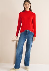 Roter Strickpullover mit hohem Kragen, kombiniert mit blauen Jeans mit weitem Bein. Ein weicher beiger Pullover wird in einer Hand gehalten. Lässiger Schuhstil.