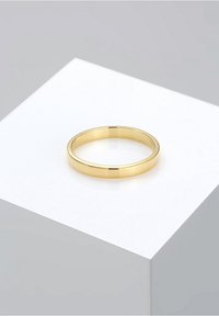 Alliance de mariage en or poli simple, présentée sur un socle blanc sur un fond gris clair.