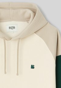 Hoodie ample beige, avec un accent vert, texture douce, capuche à cordon et un petit logo brodé sur la poitrine.