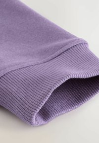 Gros plan d'une manche en tricot ribbé lavande avec des coutures visibles et une texture de tissu sur un fond neutre.