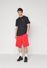 Jeune homme debout portant un t-shirt noir Nike, un short rouge Nike, des chaussettes blanches Nike et des baskets noires sur un fond clair uni.