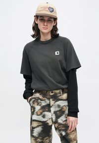 Νεαρό άτομο που φορά καπέλο μπεζ, μαύρα μπλουζάκια με ανοιχτό και σορτς μανίκι, και παντελόνι σε καφέ και γκρι χρώματα με αφηρημένο σχέδιο, που στέκεται μπροστά σε λευκό φόντο.