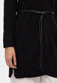 Μαύρο πλεκτό κάρντιγκαν με ριγέ μανσέτες και στρίφωμα, με μαύρη δερμάτινη ζώνη περασμένη μέσω αυτού. Η λεπτομέρεια δείχνει το χέρι που κρατά τη ζώνη.