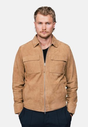 Jeune homme aux cheveux blonds courts portant une veste en daim beige avec poches avant, chemise noire et pantalon foncé, mains dans les poches, fond neutre.