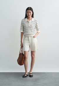 Biała polo w paski z kołnierzykiem, w zestawie z kremowymi szortami. Brązowa pleciona torba na ramię i czarne płaskie buty. Neutralne tło.