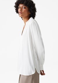 Weiße Bluse mit tiefem V-Ausschnitt und langen, voluminösen Ärmeln. Weicher Stoff mit glatter Textur, ausgestattet mit einem abgerundeten Saum für eine entspannte Passform.