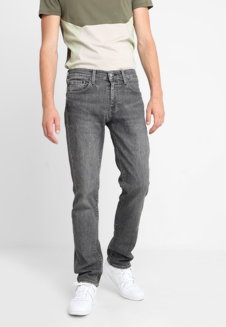 Homme portant un jean slim noir délavé, des baskets blanches et une chemise à manches courtes en bloc de couleurs vert et beige, debout contre un fond blanc.