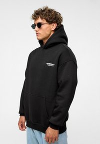 Zwarte hoodie met een voorzak en witte tekst op de linker borst. De stof lijkt zacht en textuur te hebben. Gecombineerd met blauwe jeans.