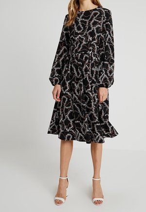 Robe midi noire avec manches longues bouffantes, motif chaîne florale abstrait blanc et rose, taille cintrée et jupe évasée, portée avec des talons blancs.