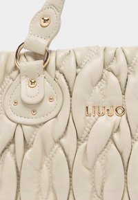 Borsa a mano in pelle beige con design trapuntato, dotata di accessori in oro, una chiusura e un logo impresso sul davanti. Texture morbida e leggero luccichio.