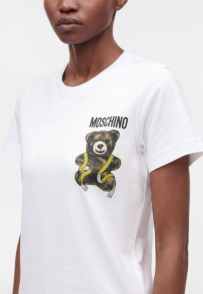Άτομο που φοράει λευκό T-shirt με το λογότυπο Moschino πάνω από μια αρκουδάκι τυλιγμένο σε κίτρινη μεζούρα, τυπωμένο στο στήθος.