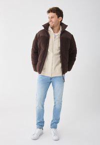 Brun fluffy jakke med en mørk brun krage, lagd over en kremfarget hettegenser. Lyseblå jeans og hvite sneakers fullfører antrekket.