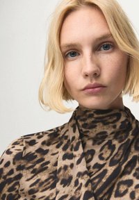 Blusa con stampa leopardata in tonalità beige e nero, caratterizzata da un collo alto e un tessuto morbido e fluente. La modella ha capelli biondi e pelle chiara.