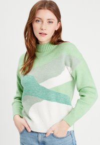 Strikket sweater i varierede nuancer af grønt og hvidt med høj hals, lange ærmer og abstrakte bølgemønstre på forsiden.