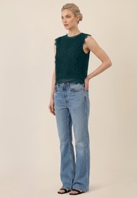 Mujer rubia con top sin mangas de encaje verde oscuro, jeans de talle alto azul claro y tacones negros abiertos, de pie con la mano en la cadera.