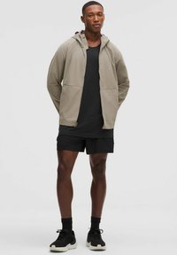 Leichte beigetönige Kapuzenjacke mit zwei Fronttaschen, getragen über einem schwarzen strukturierten Shirt und schwarzen Shorts, mit schwarzen Sneakers.